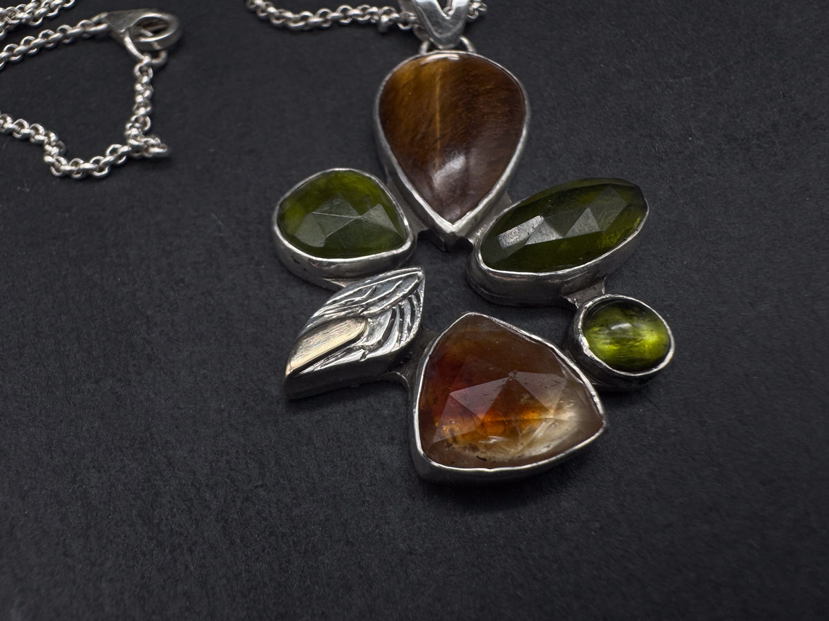 A silver wheel shaped pendant necklace featuring faceted green gemstones and a large brown tiger's eye stone, displayed on a dark textured background.