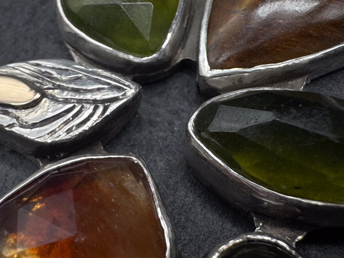 Close-up of a silver jewelry piece featuring multiple organic-shaped gemstone in olive green, amber brown, and dark brown, set against a dark textured background.