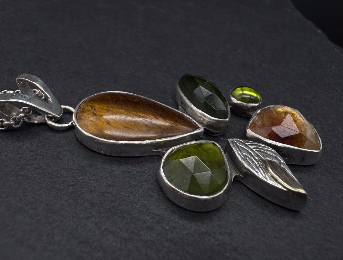 A silver pendant necklace featuring multiple teardrop and leaf-shaped gemstone settings on a dark textured background. The pendant includes a large brown tiger's eye stone, a faceted green vesuvianite stone, a smaller green stone, a citrine faceted stone, and a silver and gold leaf motif.